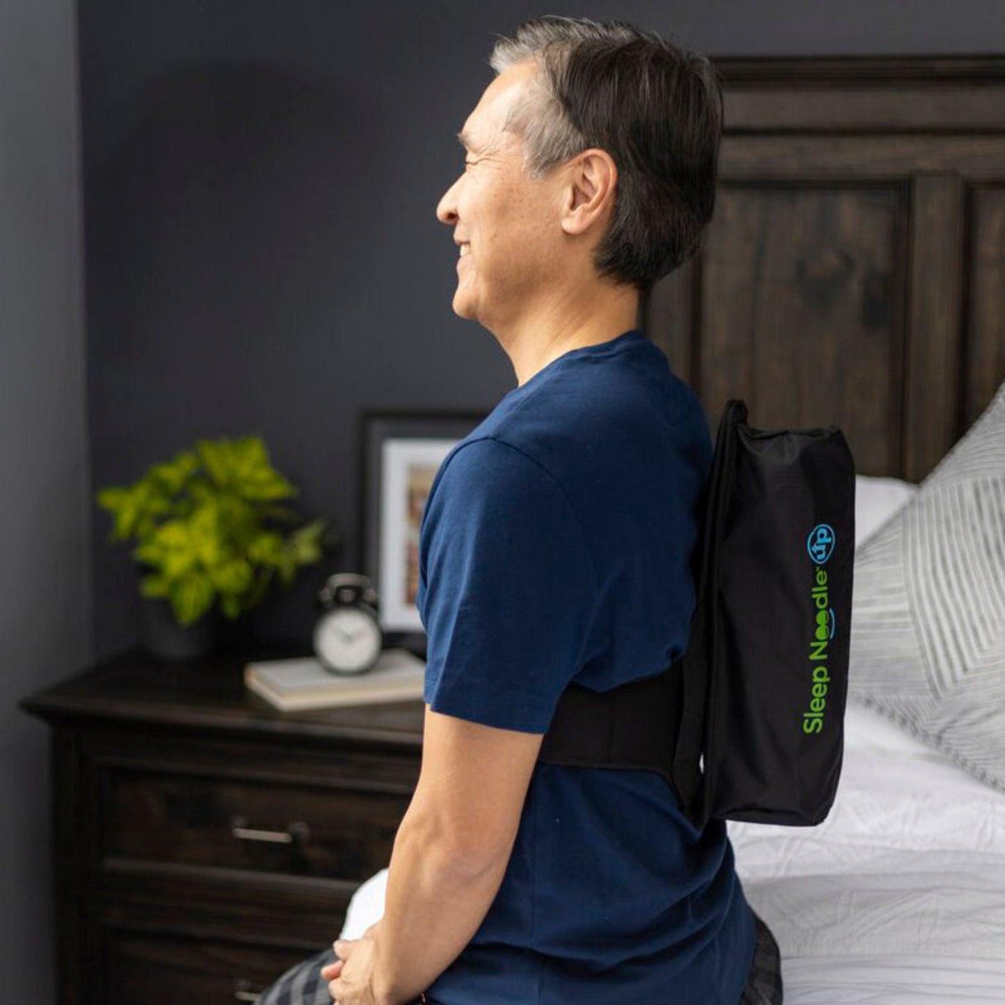 Man wearing a black back brace with 'Sleep Needler' branding in a bedroom setting.
